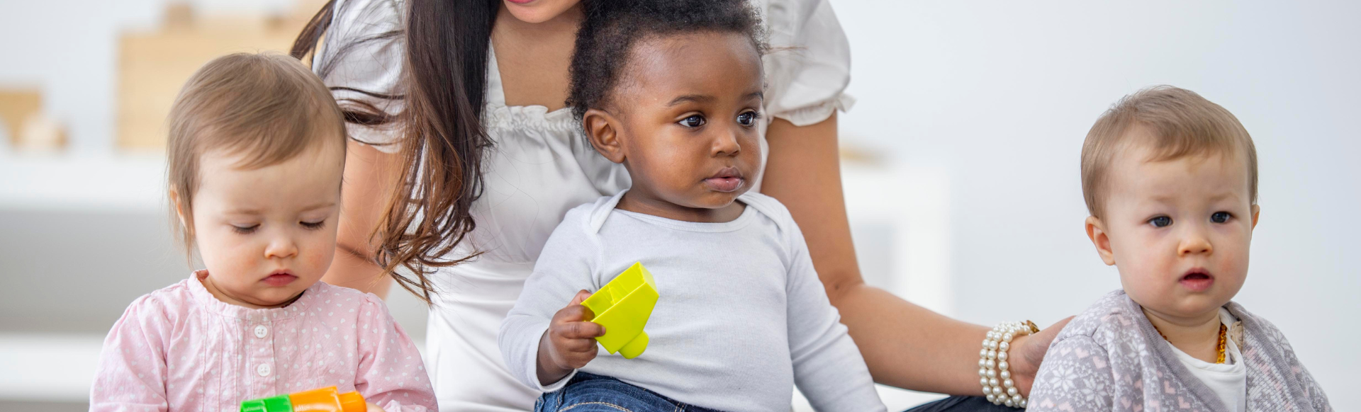 trois bambins sont assis et jouent avec des cubes de couleurs, une dame souriante se tient assise derrière eux et les surveille