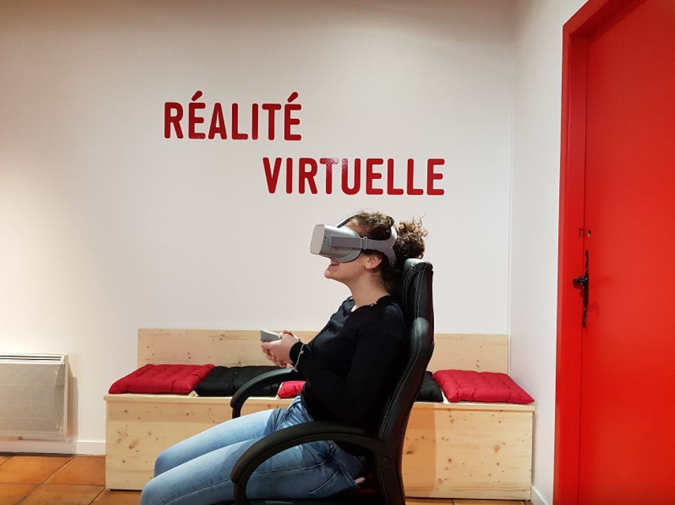 Une femme est assise sur un siège et porte un casque de réalité virtuelle. Elle sourit, au mur derrière elle il est écrit en rouge "réalité virtuelle".