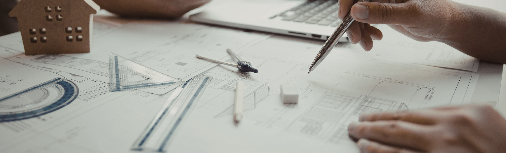 mains avec stylo, et table avec plan architecte et instruments pour dessiner (compas, équerre, règle, etc.)
