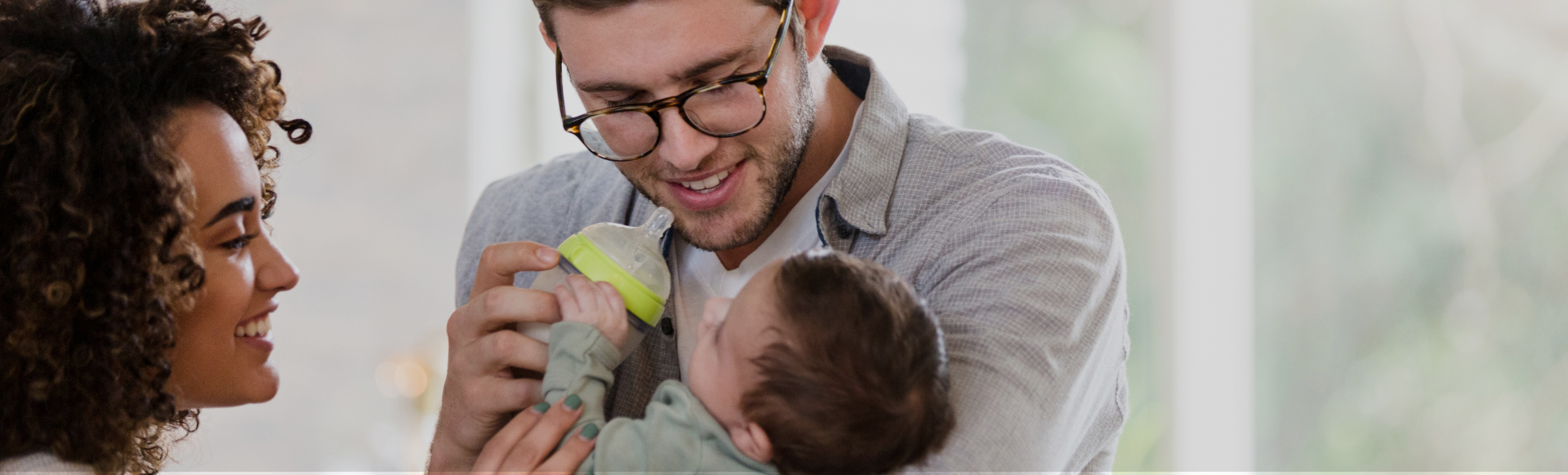 Un père tient son enfant dans les bras en souriant et va lui donner le biberon, la mère les regarde en souriant.